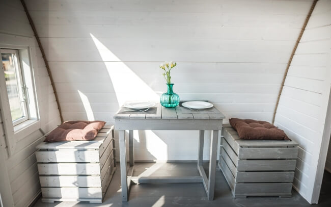 Viking Industrier 3.2 x 5.9m Camping Pod interior view of a dining setup with rustic wooden benches and table under bright natural light on a white painted wood background.