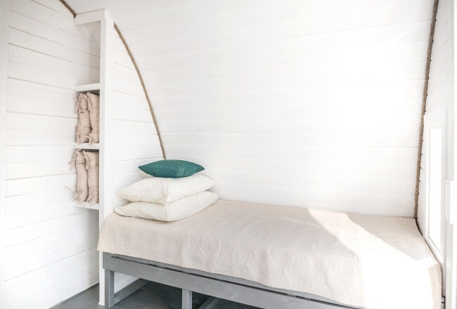 Viking Industrier 3.2 x 5.9m Camping Pod minimalist sleeping area with a single bed and open shelving, white painted wood background.