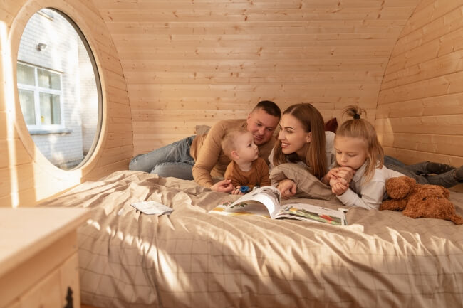 Family of four lying on a large bed in the Viking Industrier Delight Camping Pod 2.4 x 4.3m, reading a book next to a round window in a wood-paneled interior.