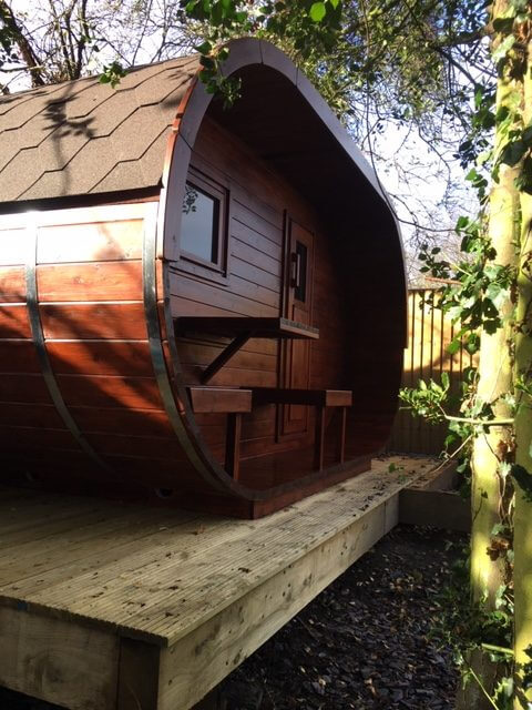 InMedias Oval Sauna installed on a raised wooden deck with deep brown wood finish, surrounded by trees  with outdoor natural background.