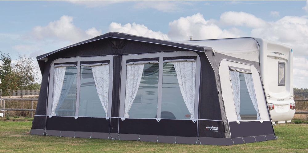Camptech Hampton DL air awning with large panoramic windows and white curtains, set up on a caravan at a campsite under a blue sky with clouds.