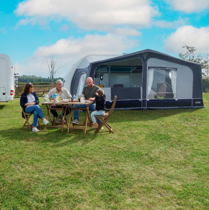 Camptech Savanna DL awning attached to a caravan, with a family dining outdoors on a grassy campsite under bright skies.