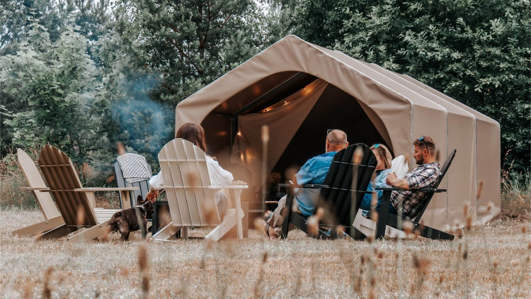 FDomes Glamping Tent with people around a fire – a tan canvas glamping tent with a group of people seated on wooden Adirondack chairs around a fire pit in a forested campsite, on a natural background.
