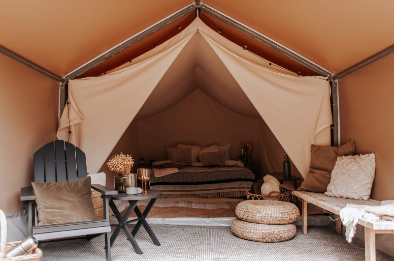 Interior view of an FDomes Glamping Tent with cozy bedding – showing a neatly styled bed, wicker poufs, and rustic decor inside a tan canvas tent, on a natural background.