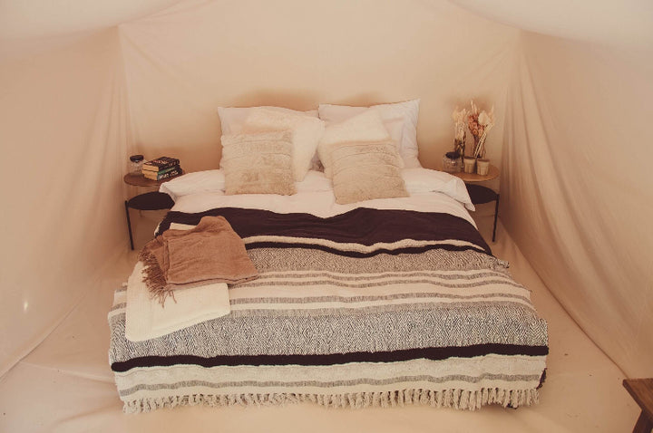 Close-up of bed inside an FDomes Glamping Tent – featuring a double bed with layered blankets and decorative pillows, flanked by small side tables, all under soft tan canvas walls, on a neutral canvas background.