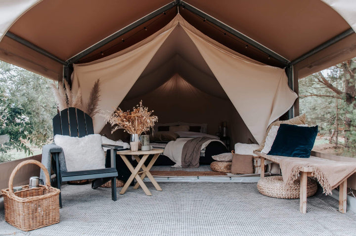 FDomes Glamping Tent with interior view of bed and seating area – showing a black chair with pillow, bench seating, wicker poufs, and a made-up bed in the background under a peaked tan canvas roof, on a natural background.