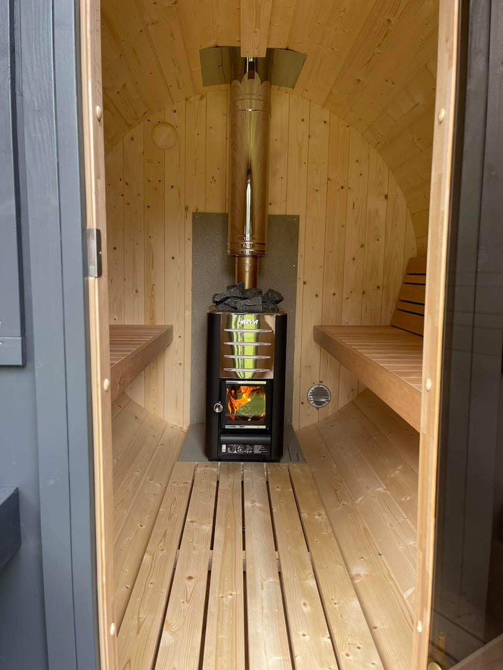 Interior view of InMedias Barrel Sauna 280 showing wooden benches on both sides and a central Harvia stove – taken from doorway with natural light.