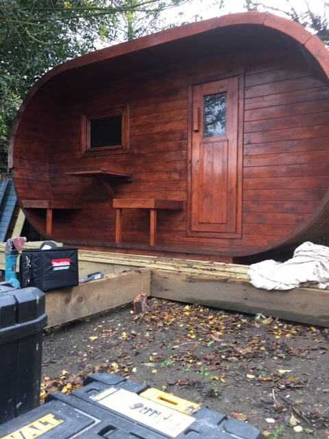 InMedias Oval Sauna with deep red-stained wood under construction, tools scattered in front with outdoor background and leaves.