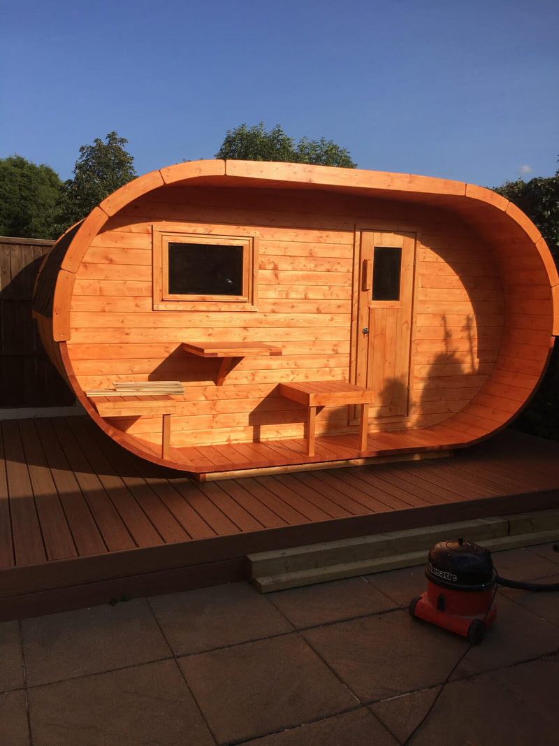 InMedias Oval Sauna glowing in sunset light on a deck, warm-toned wood finish with  outdoor background with shadows on wooden fence.