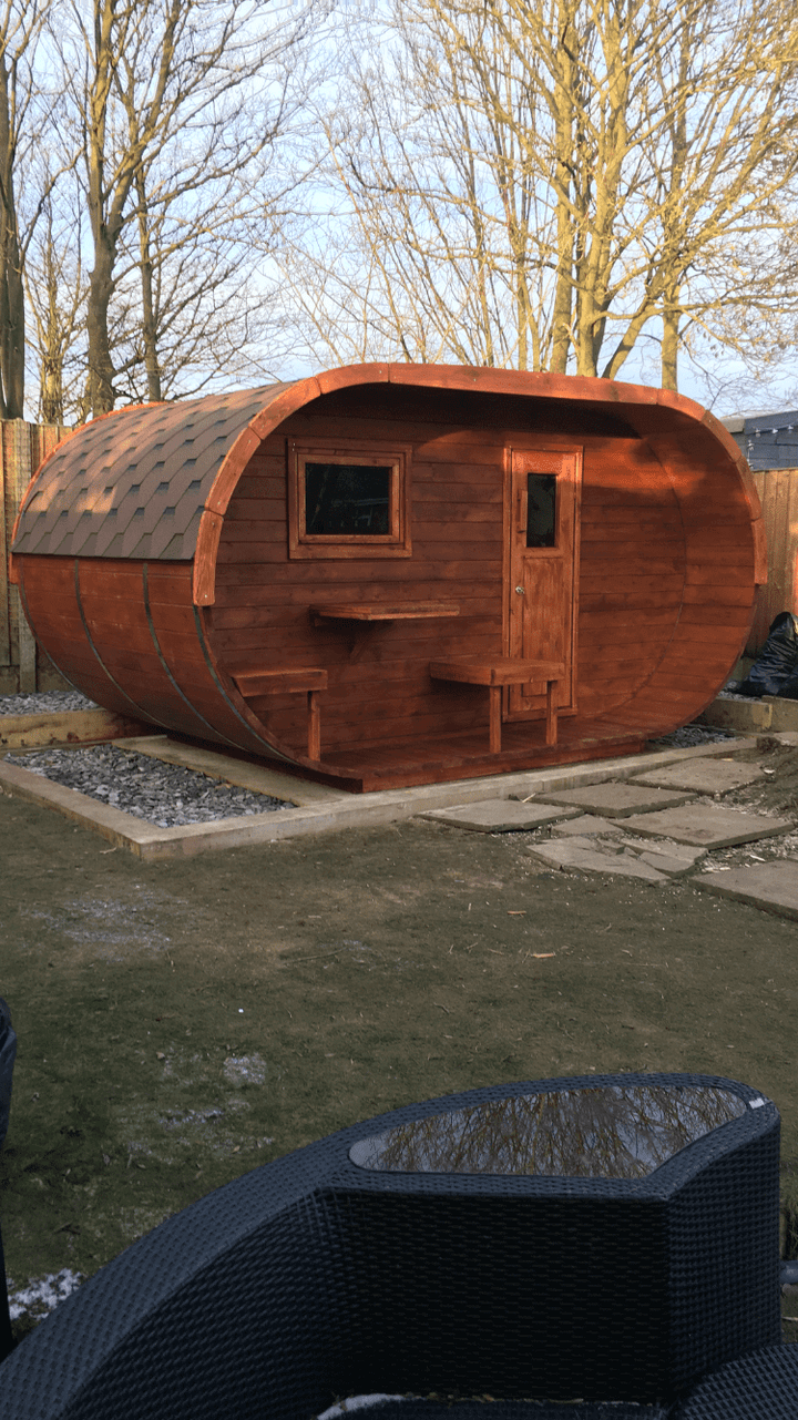 InMedias Oval Sauna with deep cherry-stained wood finish and black shingle roof with  backyard background with leafless trees.
