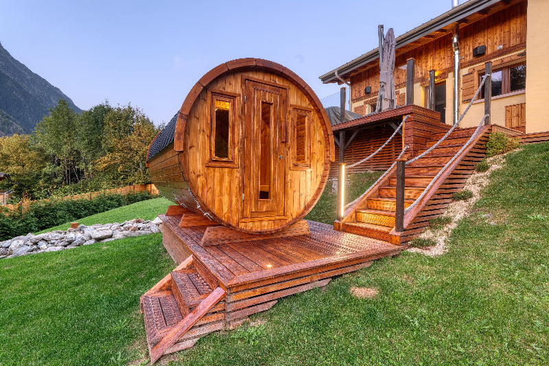 InMedias Barrel Sauna 400 Deluxe situated on an elevated wooden platform beside a rustic home in a mountainous landscape, on a blue sky background