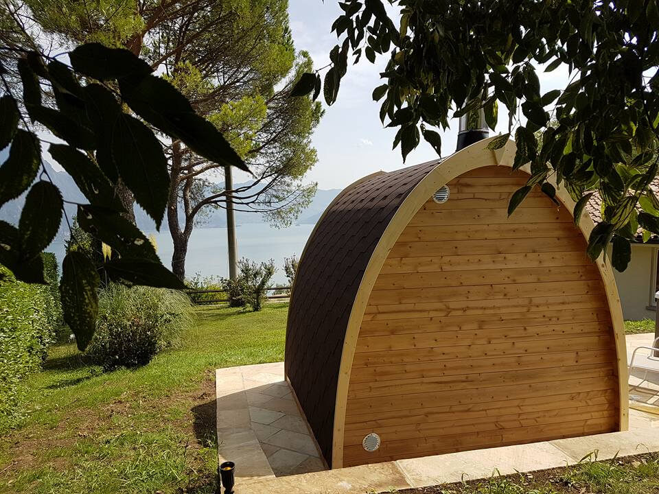 Rear view of InMedias Sauna Pod 300 showing wooden paneling and vent ports on a sunny day with tree shadows and a lake view.