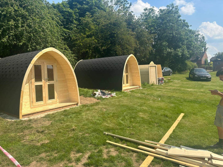 Outdoor view of two InMedias Camping Pod 480 units being installed on a grassy field with tools and materials nearby.