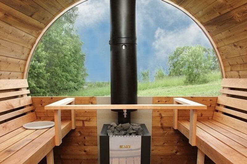 Interior view of InMedias Barrel Sauna 400 with wooden benches and a black Harvia stove, showing a curved glass wall overlooking a grassy landscape with a natural background.