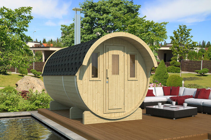 InMedias Barrel Sauna 330 in light wood finish placed on a deck beside red and white outdoor furniture — on a garden background.