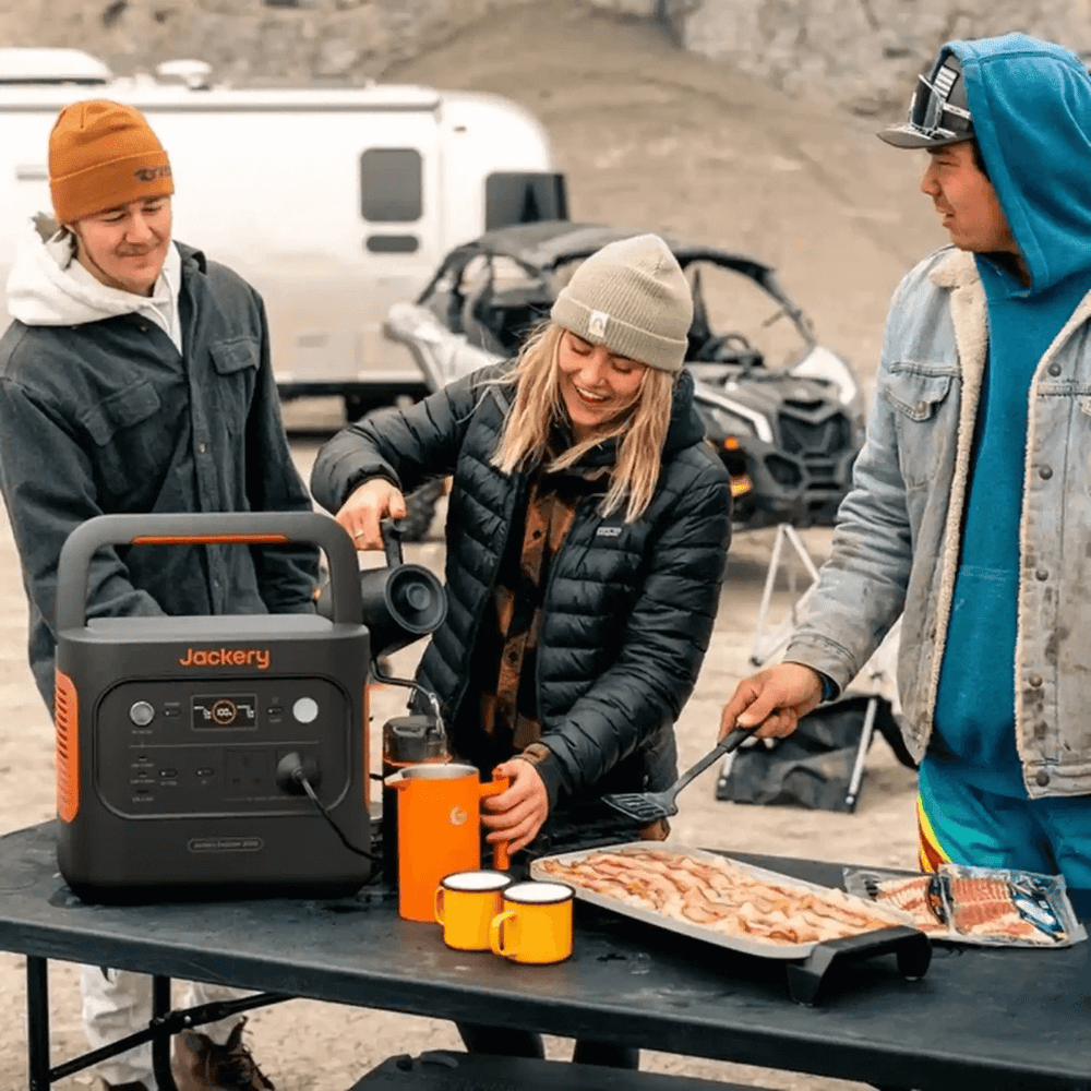 Jackery Explorer 2000 V2 portable power station powering outdoor cooking equipment at a campsite with three friends in jackets, on a cloudy background.