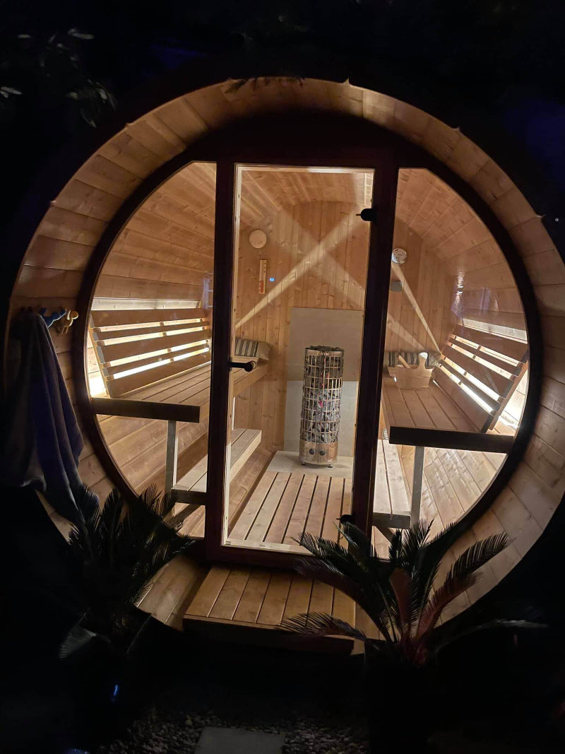 Nighttime view of InMedias Barrel Sauna 280 Deluxe lit from inside, showing benches and heater through a large glass door