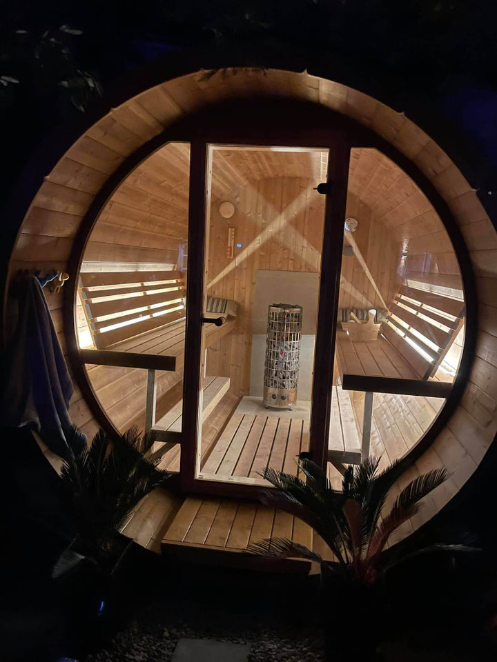 Nighttime view of InMedias Barrel Sauna 280 Deluxe lit from inside, showing benches and heater through a large glass door