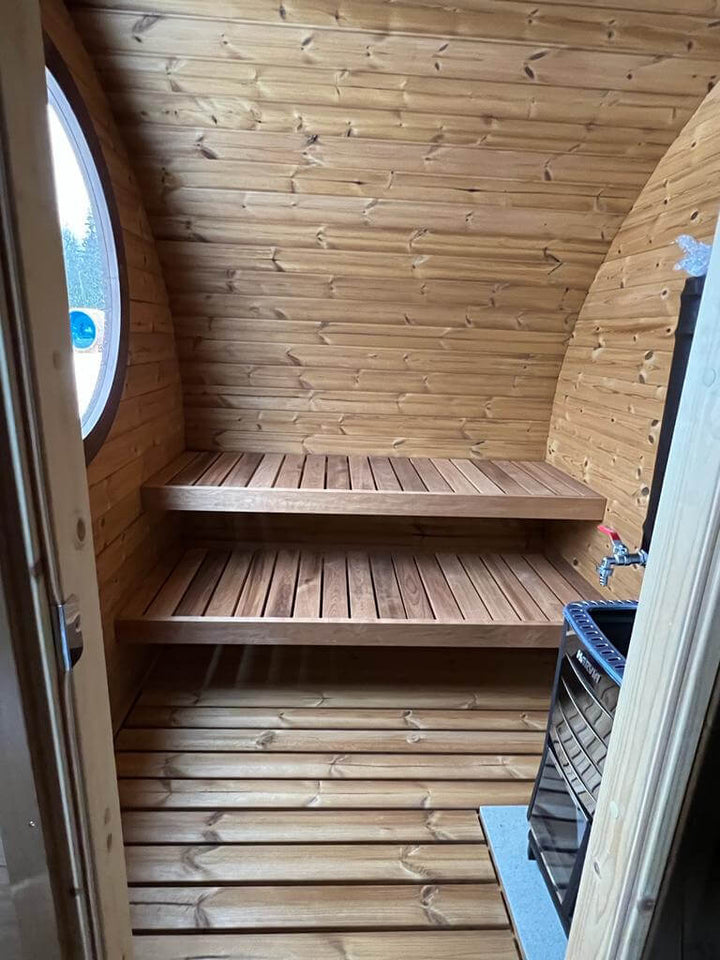 Interior shot of the InMedias Oval Sauna Frodo 195 showing wooden benches and a heater, with a round window on the left, photographed in natural daylight.