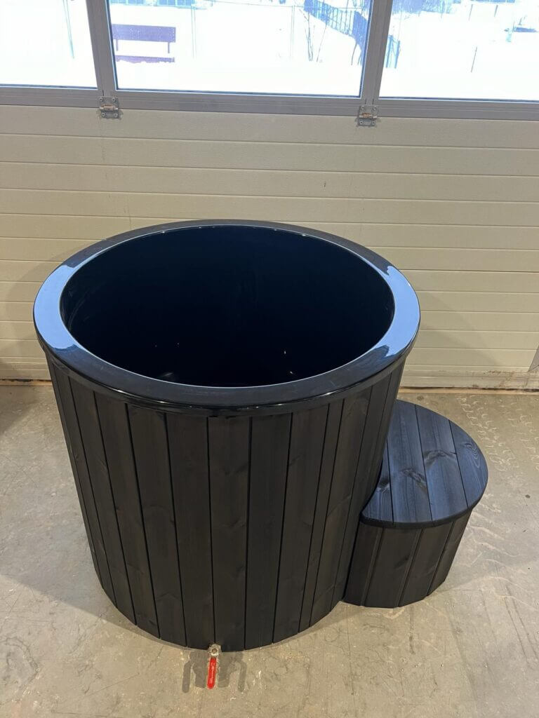 Black wooden cold tub with glossy black interior and rounded step, placed indoors near large windows with natural light.