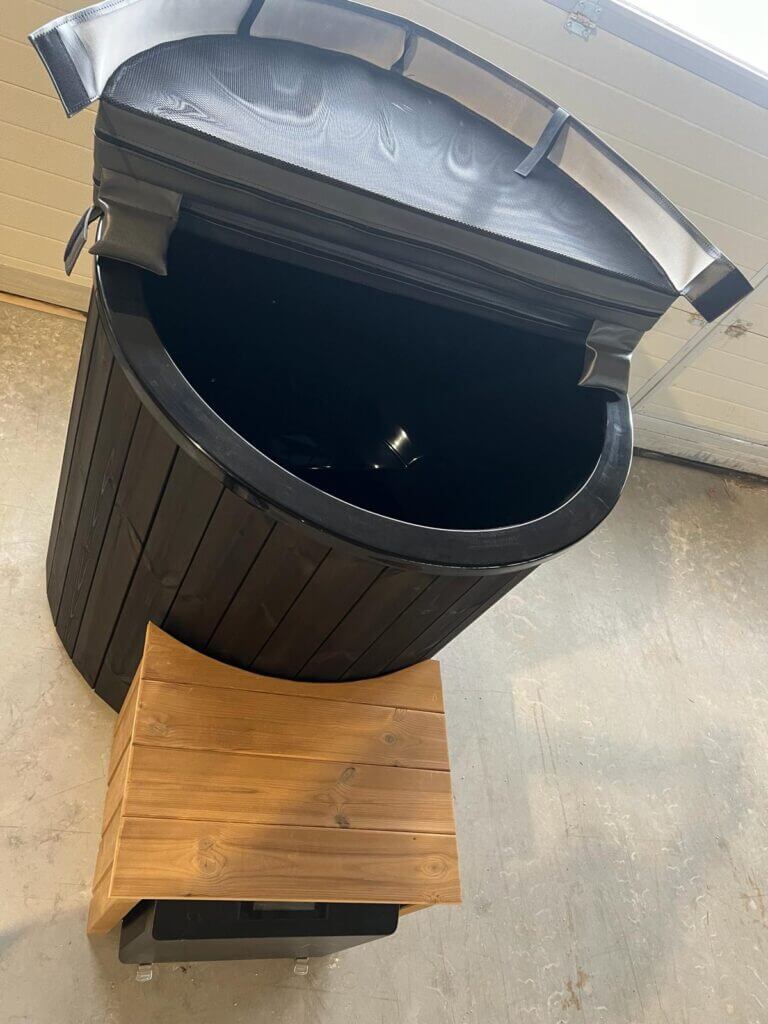 Black wooden cold tub with partially opened insulated lid and a small wooden box step, indoors on a light concrete floor.