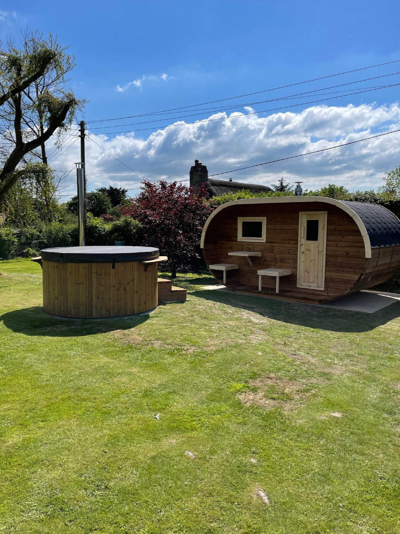 InMedias Oval Sauna placed beside a circular hot tub on grass with a stone pathway  with garden background with blue sky and trees.