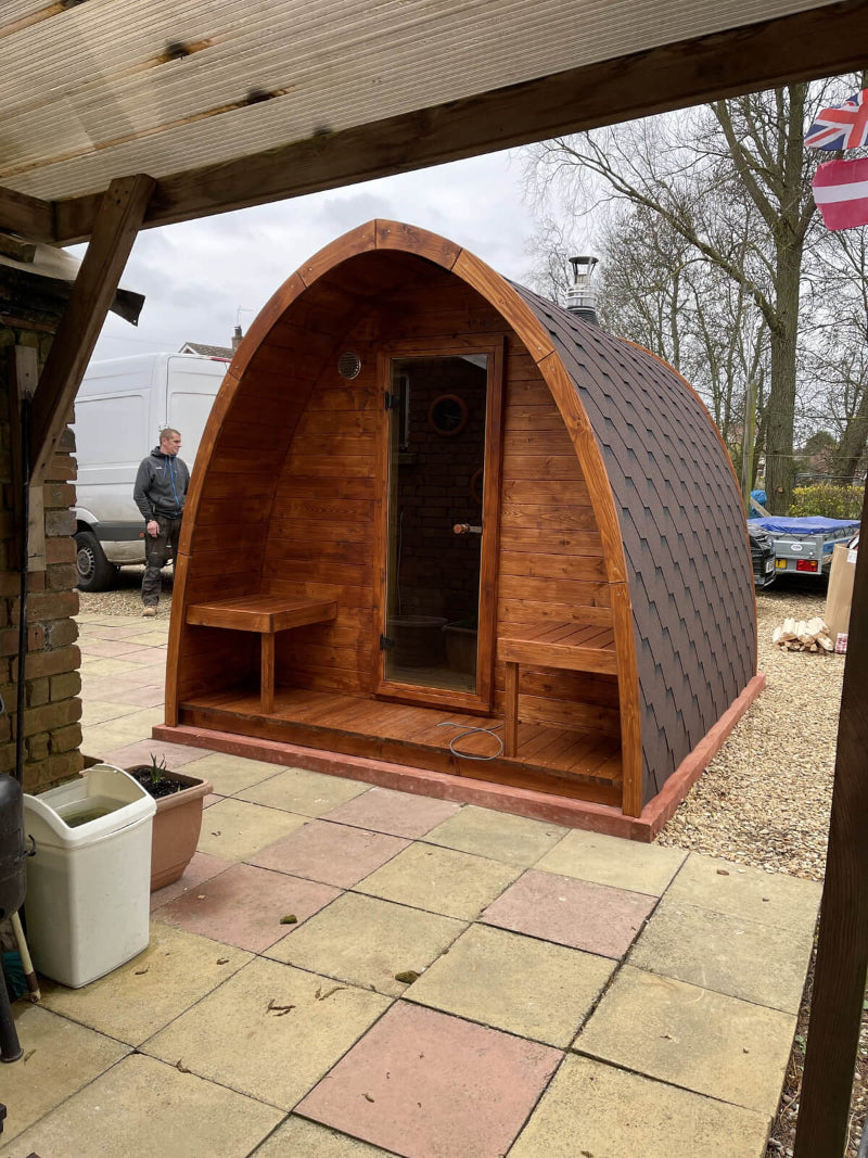 InMedias Sauna Pod 300 with rich brown finish and shingle roof located on multicolored tiled patio under a covered wooden frame structure.