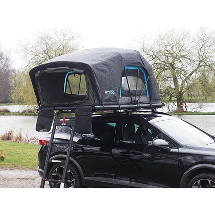 Skypod Lite rooftop tent mounted on a black SUV parked beside a lake in a countryside campsite.