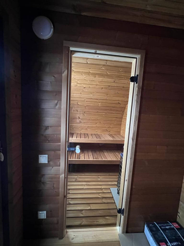 View through the doorway into the sauna room of the InMedias Oval Sauna Frodo 195, showing dark wooden walls and a heater on the right, indoor lighting.