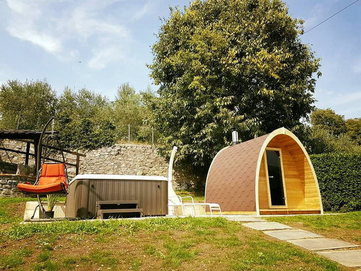 InMedias Sauna Pod 300 beside a hot tub and lounge chairs in a backyard garden on a sunny day with blue sky and trees.