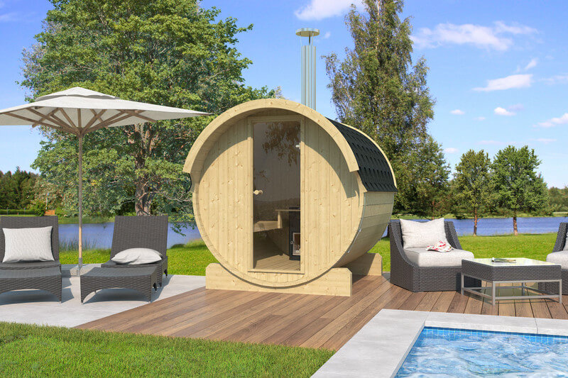 A cylindrical InMedias Barrel Sauna 220 positioned on a wooden poolside deck with lounge chairs and umbrella, surrounded by grass and trees under a blue sky.