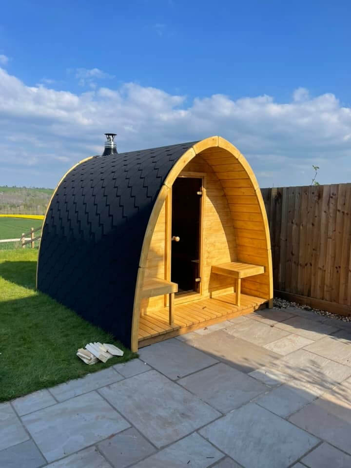 InMedias Sauna Pod 300 with light wood body and black roof placed in a grassy backyard with wooden fencing on a bright blue sky background.