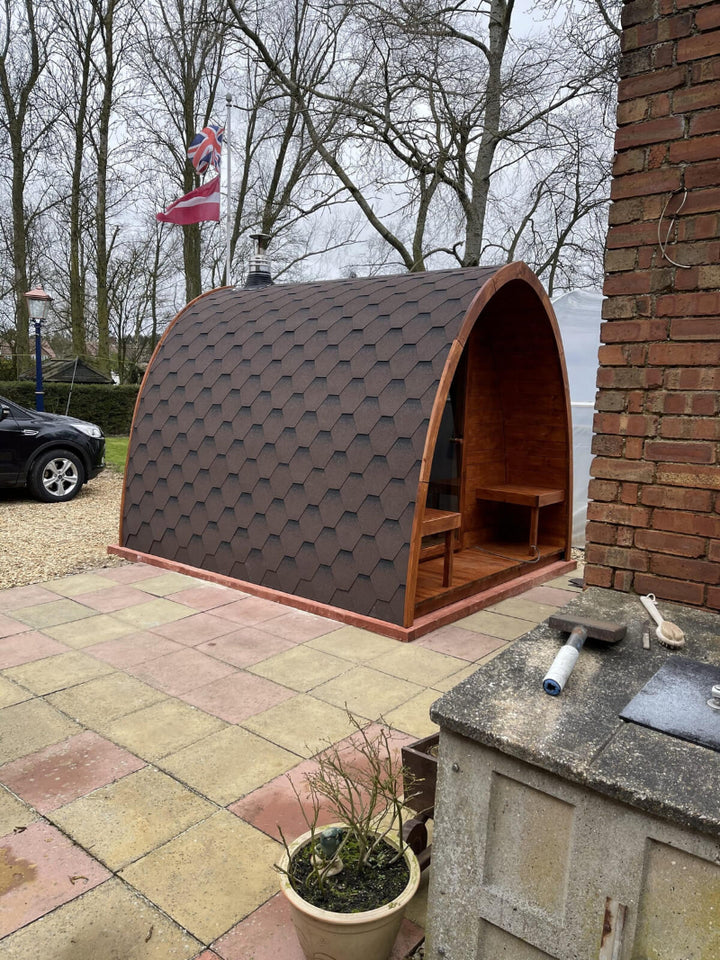 InMedias Sauna Pod 300 with hexagonal dark shingle roof positioned on paved tiles beside a brick wall on a cloudy gray sky background.