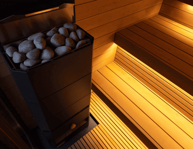Detailed view of the black sauna heater filled with sauna stones, beside illuminated wooden bench seating inside the Saunum AirCube One.