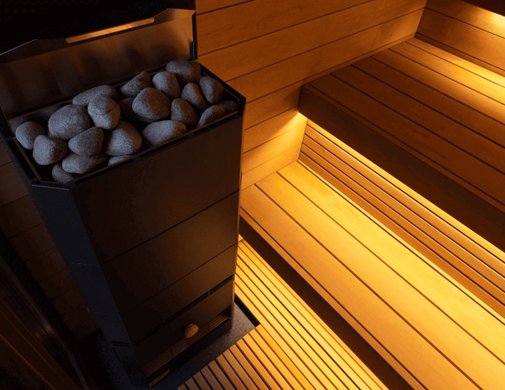 Detailed view of the black sauna heater filled with sauna stones, beside illuminated wooden bench seating inside the Saunum AirCube One.