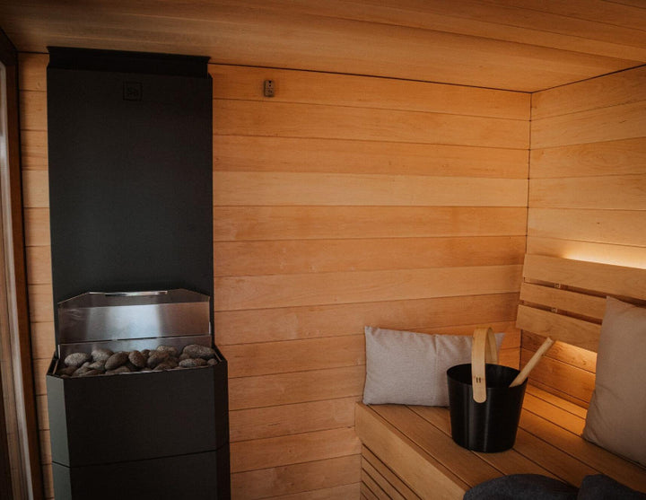 Inside the Saunum AirCube Terrace sauna featuring a black stone heater, wooden walls, seating with cushions, and a traditional sauna bucket and ladle.