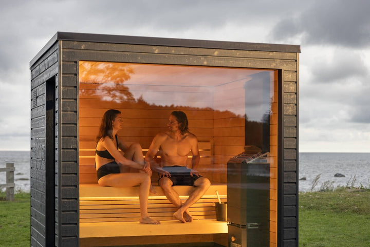 A couple relaxing inside a modern black wood-clad sauna with a full glass front, set on a grassy field near the ocean.