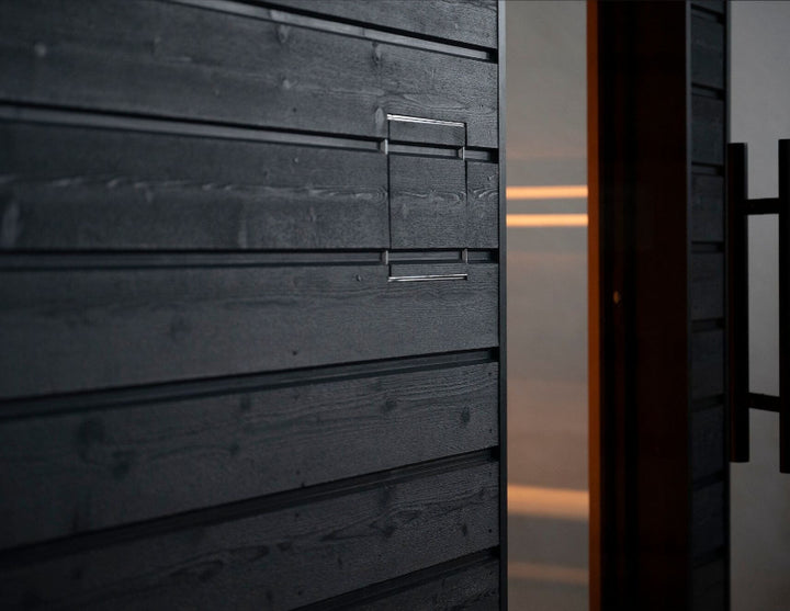 Close-up of a matte black wooden sauna exterior panel with subtle handle detailing and soft reflections.