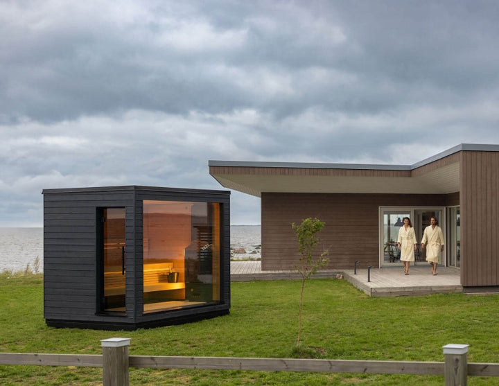 Saunum Classic sauna placed on a grassy lawn beside a modern home, overlooking a seafront with cloudy skies.