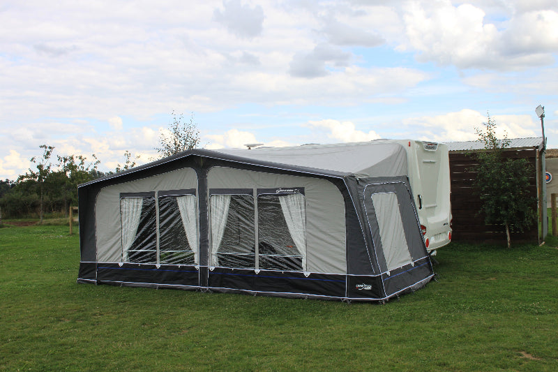 Camptech Savanna DL full-size caravan awning with clear front windows and white curtains, attached to a caravan on a green campsite under a cloudy blue sky.