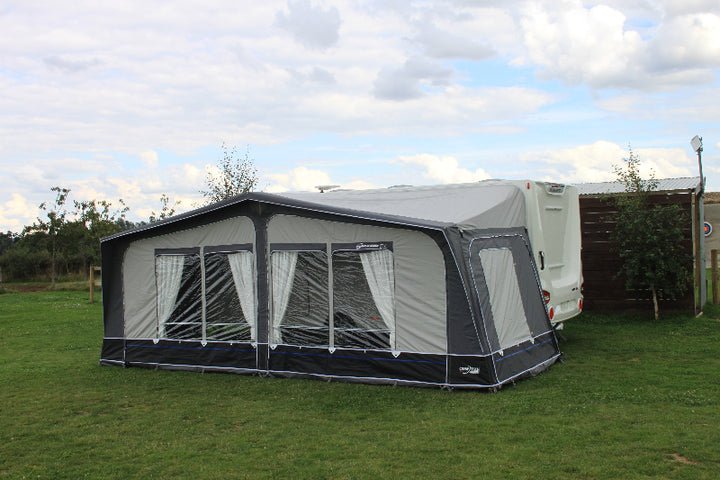 Camptech Savanna DL full-size caravan awning with clear front windows and white curtains, attached to a caravan on a green campsite under a cloudy blue sky.