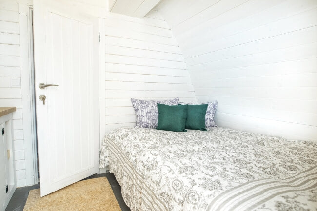 Viking Industrier 3.2 x 5.9m Camping Pod cozy sleeping area with patterned bedding and decorative pillows against white panel walls on a white wood background.