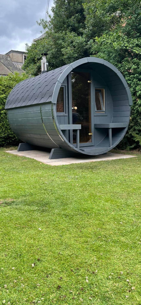 InMedias Barrel Sauna 280 in matte gray finish with glass door and twin benches, placed on a patio – photographed outdoors with trees and cloudy sky.