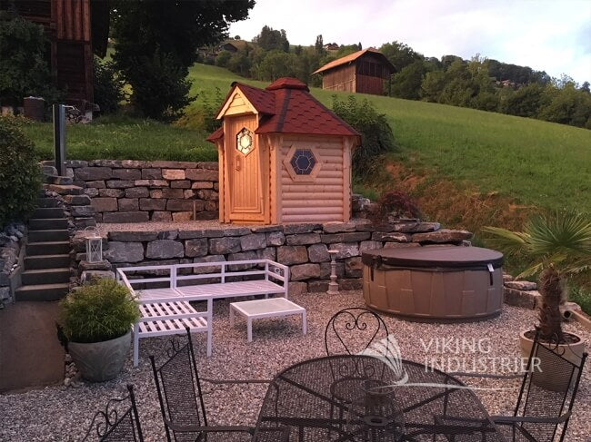 Viking Industrier sauna cabin 4.5m² installed in a landscaped garden with stone wall, hot tub, and outdoor furniture, surrounded by hills and houses in the distance.