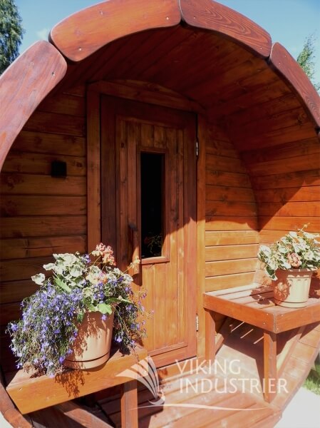 Front view of a Viking Industrier Barrel Sauna 1.9 x 2.5m with wooden door and built-in bench seating on both sides, surrounded by potted flowers, outdoor setting with trees.