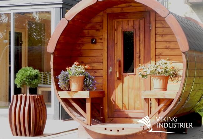 Full front view of the Viking Industrier Barrel Sauna 1.9 x 2.5m showcasing its circular wood design, symmetrical bench seating with potted plants, and a central vertical window on the door.
