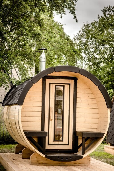 Front view of the Viking Industrier Barrel Sauna 1.9 x 3m with terrace, featuring a natural wood finish and black roof shingles, placed outdoors with green trees in the background.