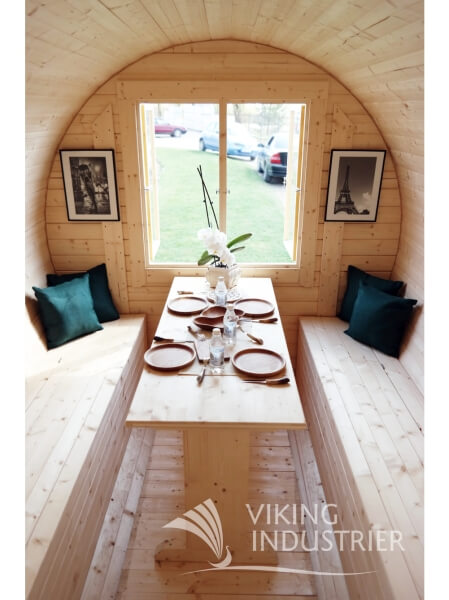 Interior of Viking Industrier Barrel Sauna 2.2 x 5.9m featuring a dining setup with wooden benches and table, decorative pillows, and framed pictures, with a green outdoor view through a window.