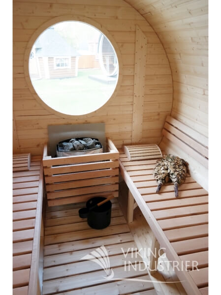 Interior view of Viking Industrier Barrel Sauna 2.2 x 5.9m showing a circular window, wooden benches, sauna heater, and sauna accessories including a bucket and birch branches.