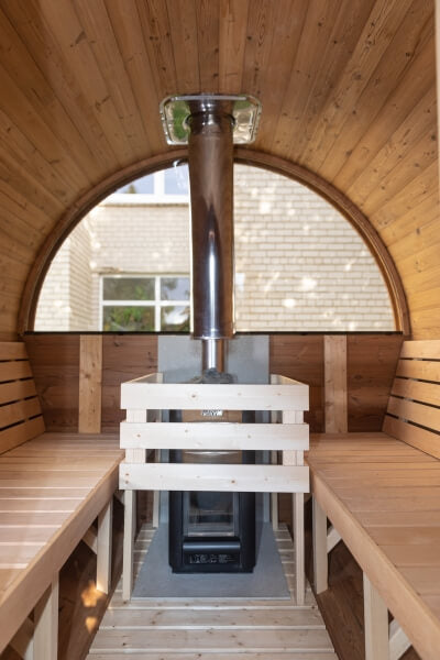 Interior view of a Viking Industrier Barrel Sauna with a wood-burning stove and curved wooden benches, with a brick wall seen through the back window.